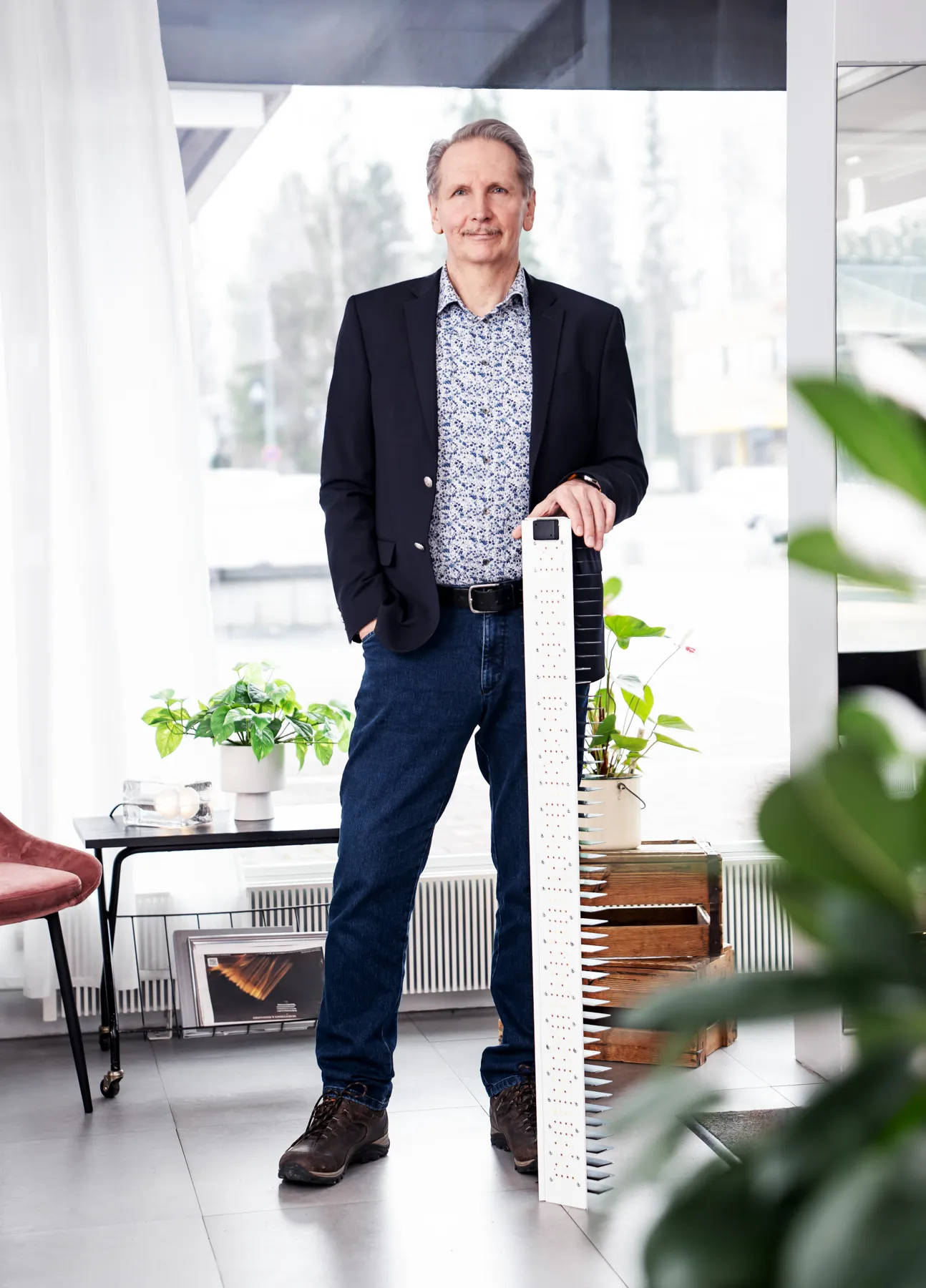 Keskellä huonetta seisoo mies tummassa bleiserissä, sinisissä farkuissa ja ruskeissa kengissä, pitäen kädessään valkoista, rei'itettyä ja sahalaitaista esinettä. Taustalla näkyy ikkuna, kasveja ja huonekaluja. Valoisa ja moderni sisustus.